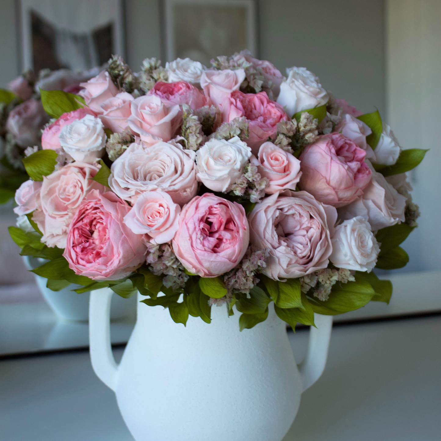 A white vase filled with pink and white roses and greenery on a table.