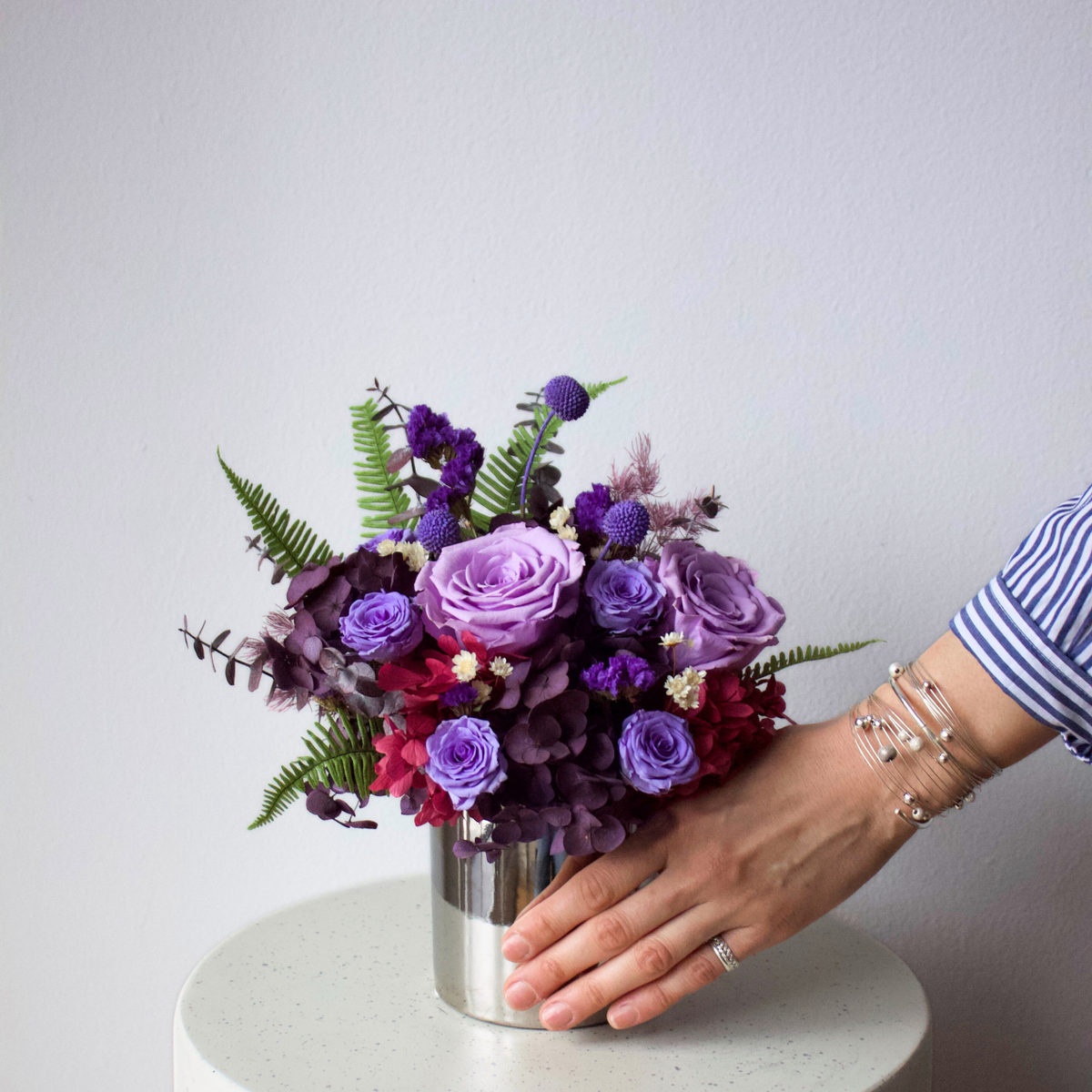 A hand arranges Eos Blooms' Preserved Flower Subscription in a silver vase—timeless beauty.