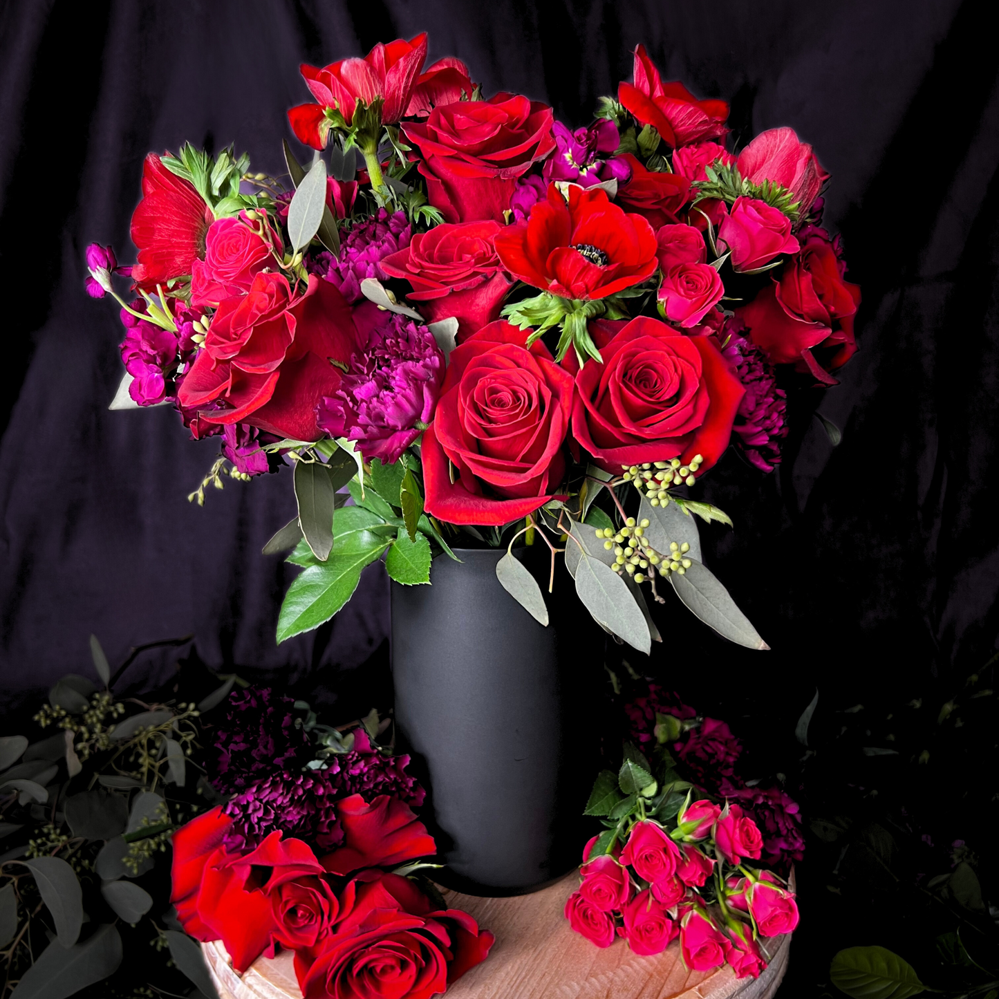 The Bisous by Eos Blooms: red roses in a black vase on wood with a dark background.
