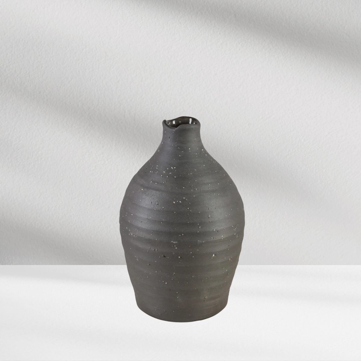 A black Eos Blooms Tikal Vase with a small spout sits on a white surface against a light background.