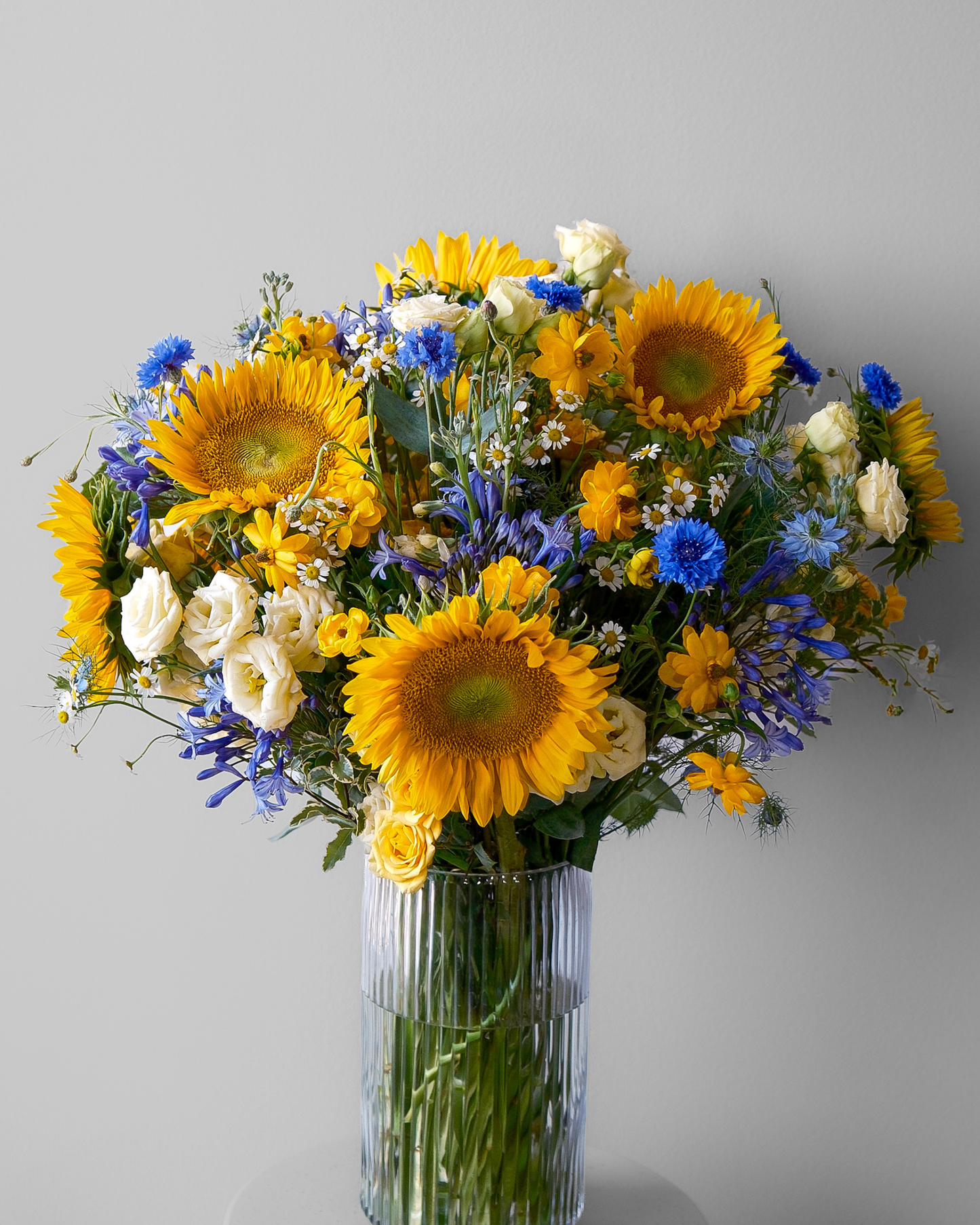 Eos Blooms’ The Starry Night: sunflowers and blue wildflowers in a vase on a gray backdrop.