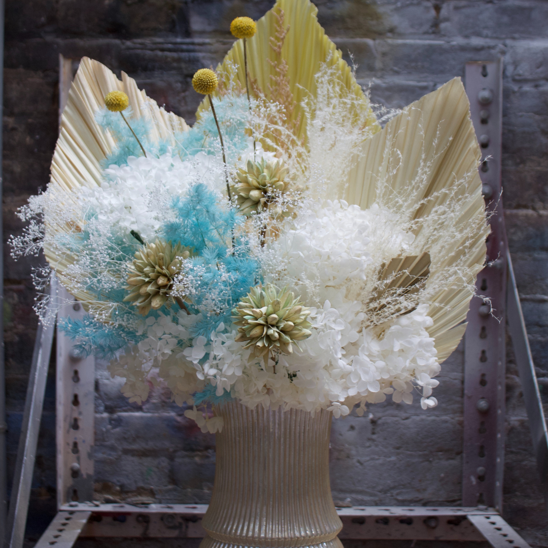 Decorative floral arrangement with large leaves and small flowers in a textured vase against a stone wall.