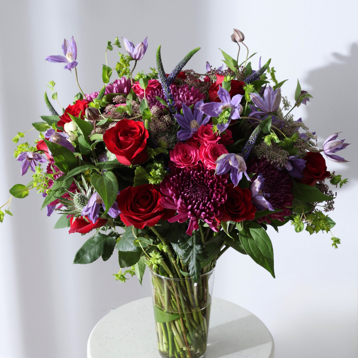 Bouquet of red roses and purple flowers in a glass vase on a white table.
