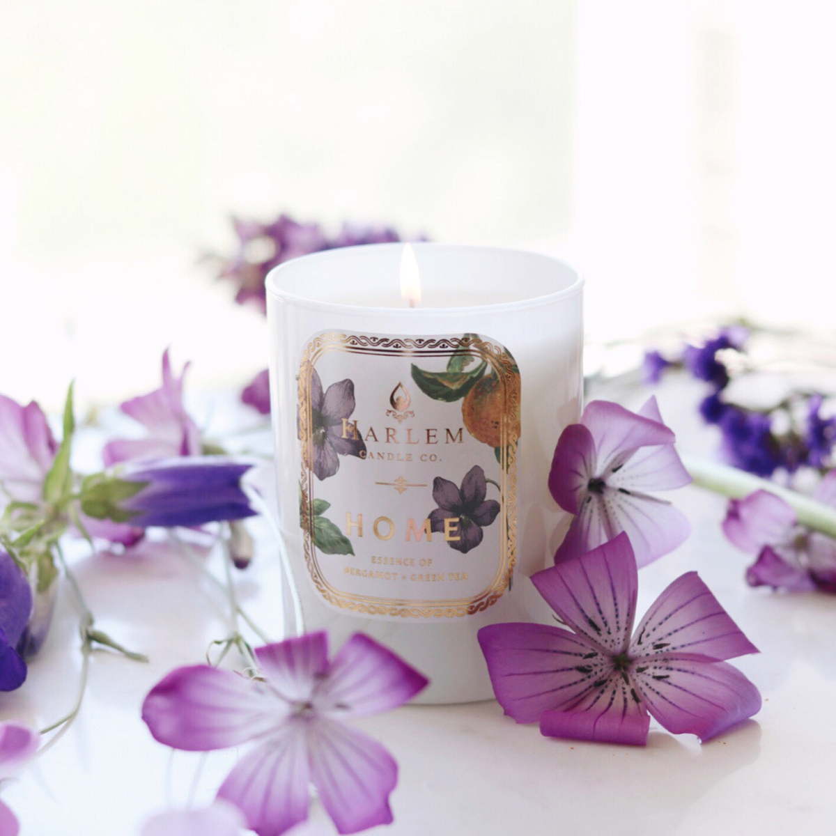 A lit Harlem candle surrounded by scattered purple flowers on a white surface.
