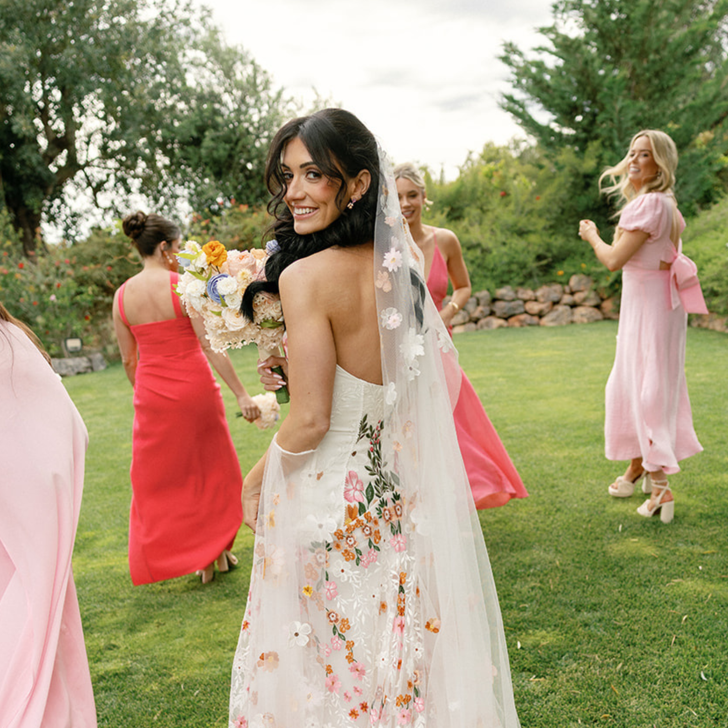 A bride in "The Promise" by Eos Blooms smiles, with bridesmaids in pink dresses nearby.