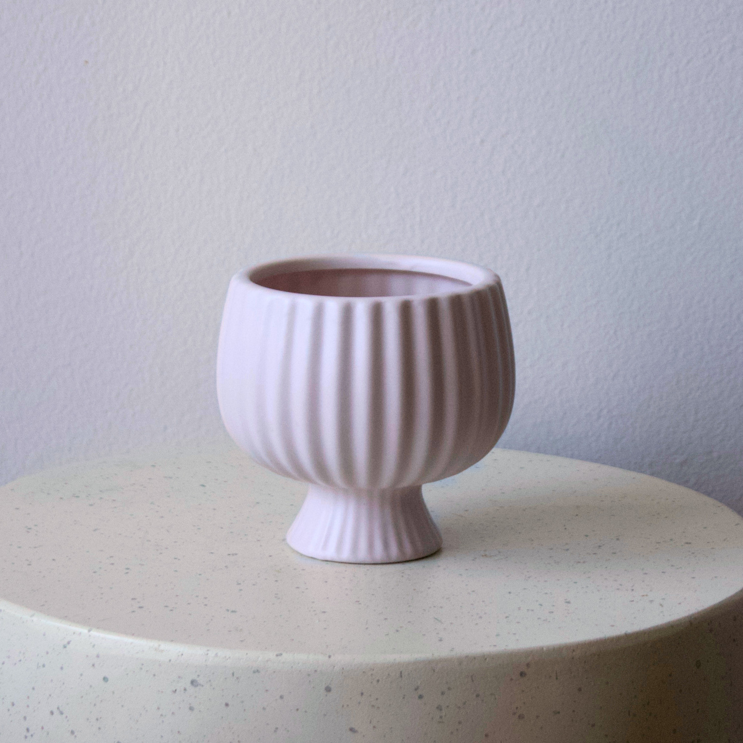 The Eos Blooms Small Blush Pedestal rests on a speckled surface beside a pale wall and vase.