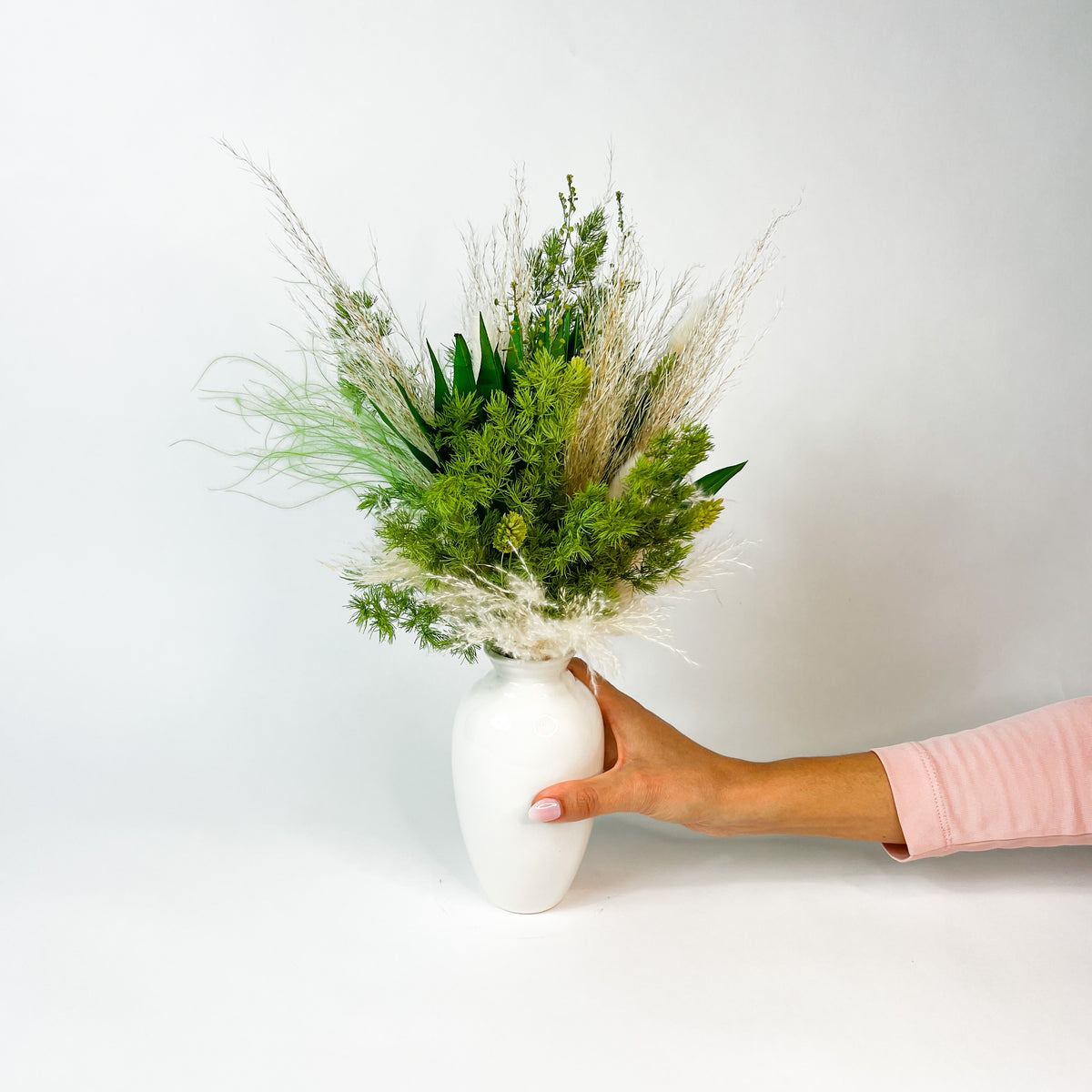 A hand holds Eos Blooms' Botanical—a white vase with preserved flowers—against a white background.