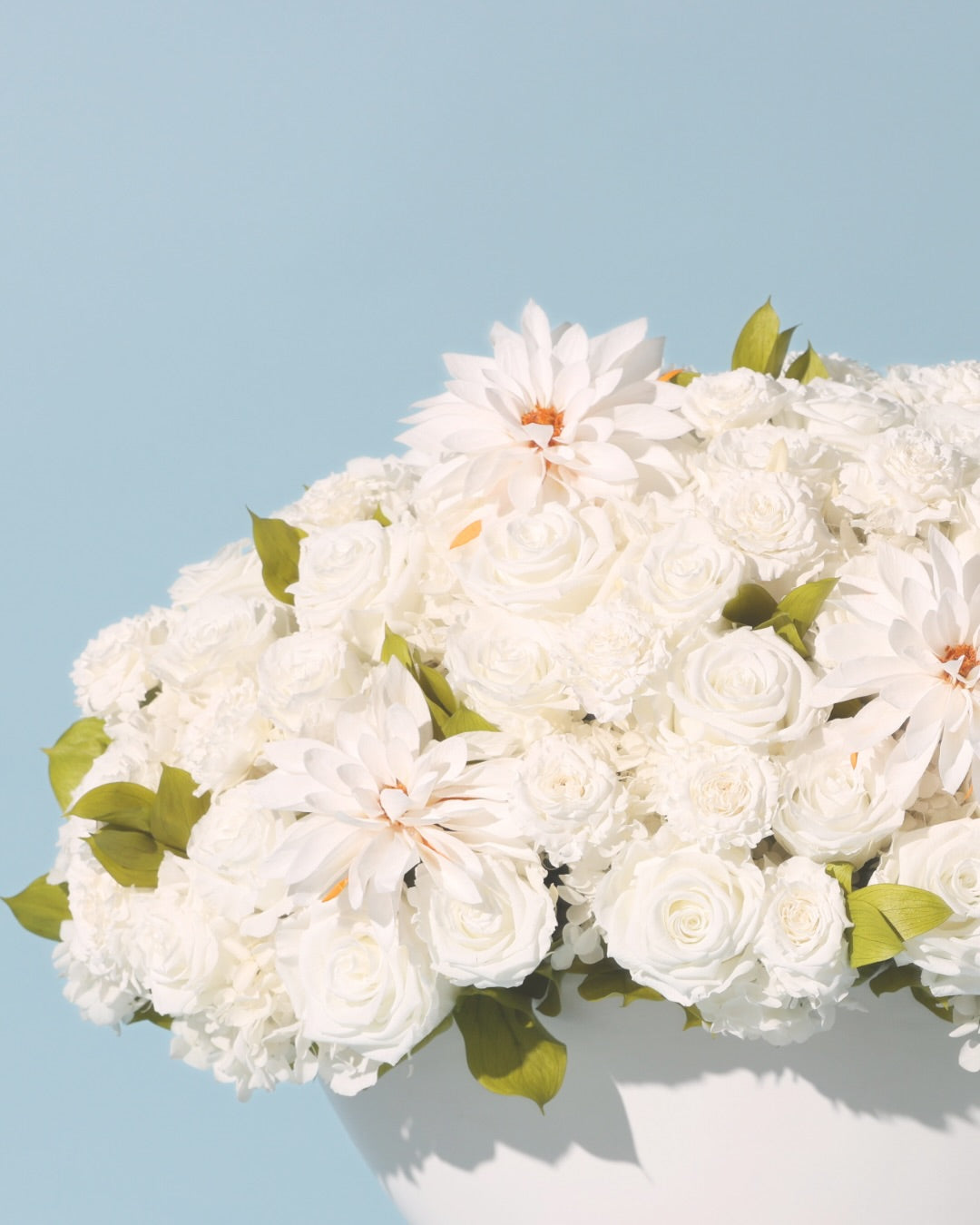A bouquet of white flowers, including roses and dahlias, with green leaves in a white vase, set against a light blue background.