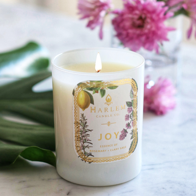 Lit Joy candle in a white glass jar with flowers and leaves on a marble surface.