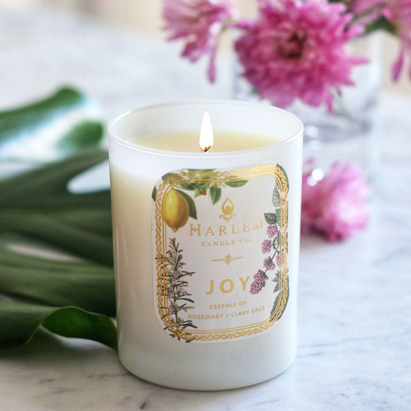 Lit Joy candle in a white glass jar with flowers and leaves on a marble surface.