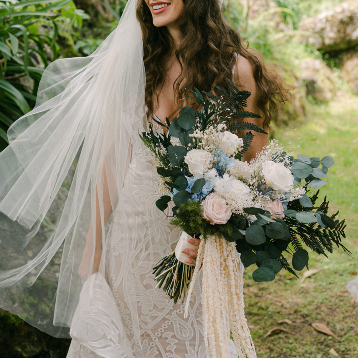 Bride outdoors in lace with The Promise bouquet by Eos Blooms, smiling amid florals and greenery.