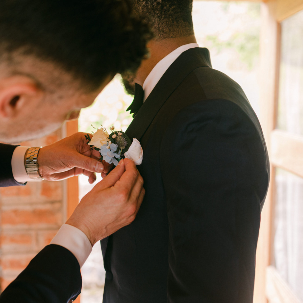 A man fastens Eos Blooms' The Promise boutonnière on another's suit near a sunlit doorway.
