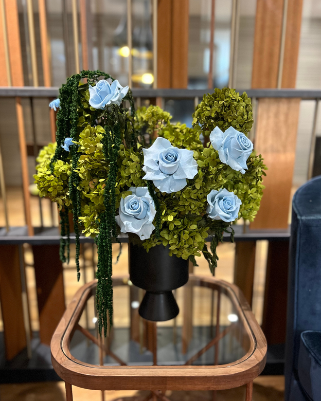 A black vase with blue roses and green foliage on a glass-top wooden table.