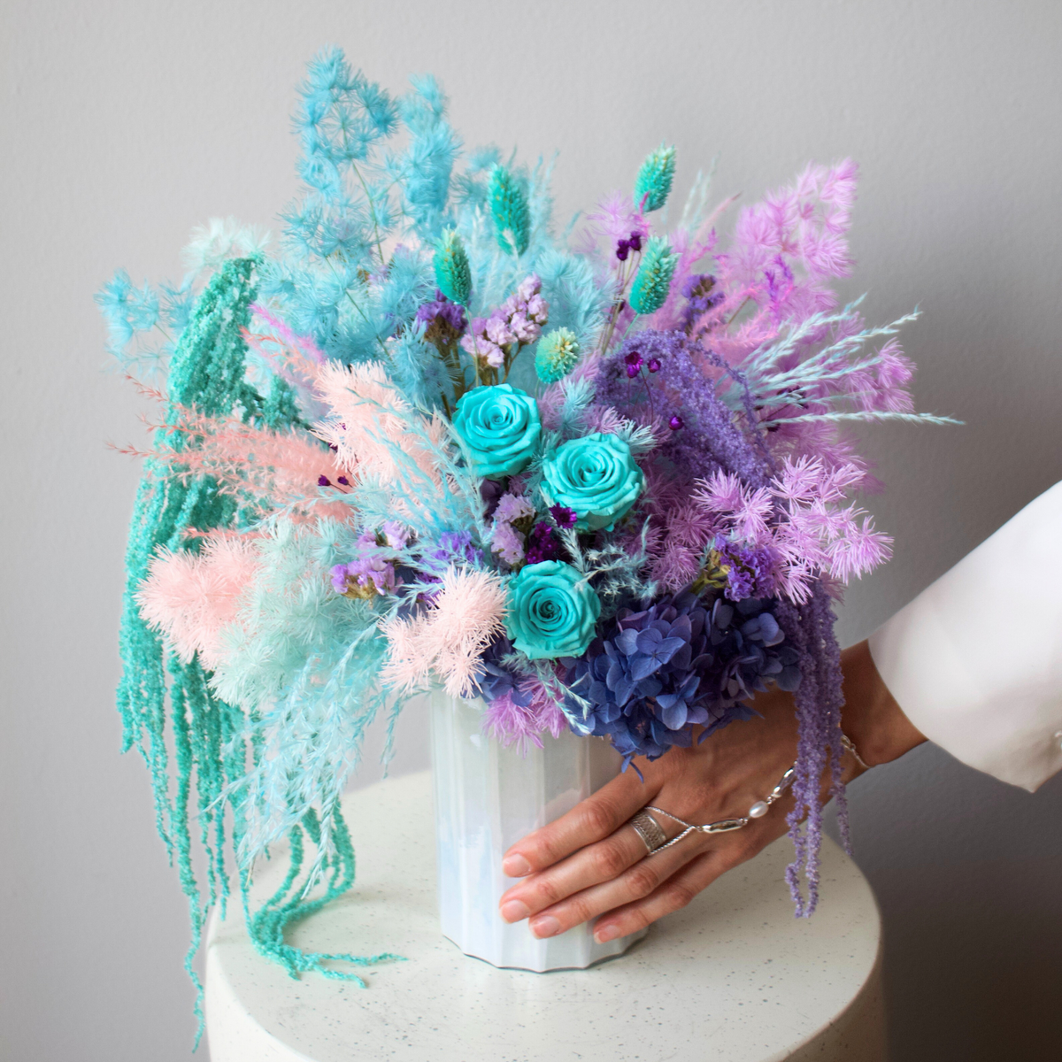 A hand touches Eos Blooms' La Sirena preserved floral arrangement on a white round table.