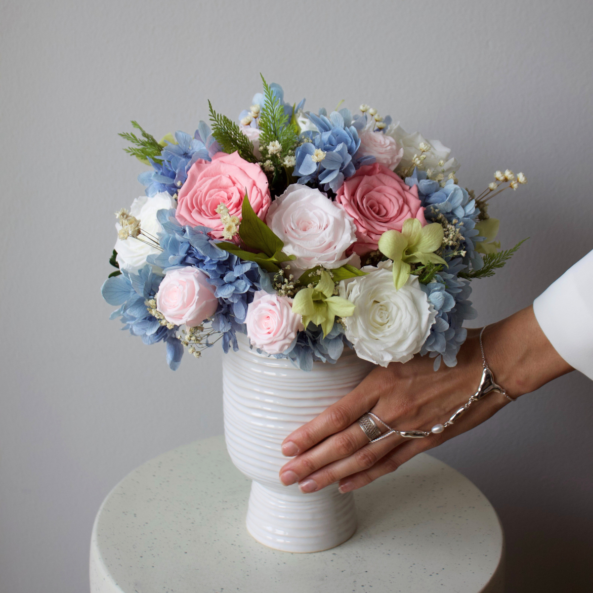 A hand touches an Eos Blooms Preserved Flower Subscription on a round white table.