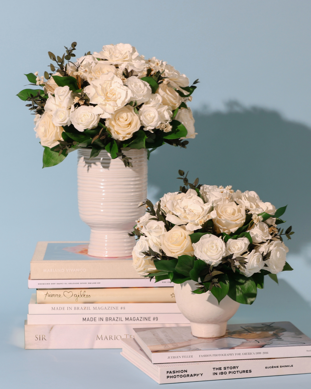 Two Eos Blooms Snowdrop vases with Forever Roses on books, set against a light blue background.