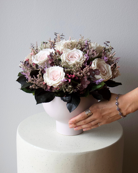 Hand adjusting The Chantilly by Eos Blooms, a pink rose and purple bouquet, on white pedestal.