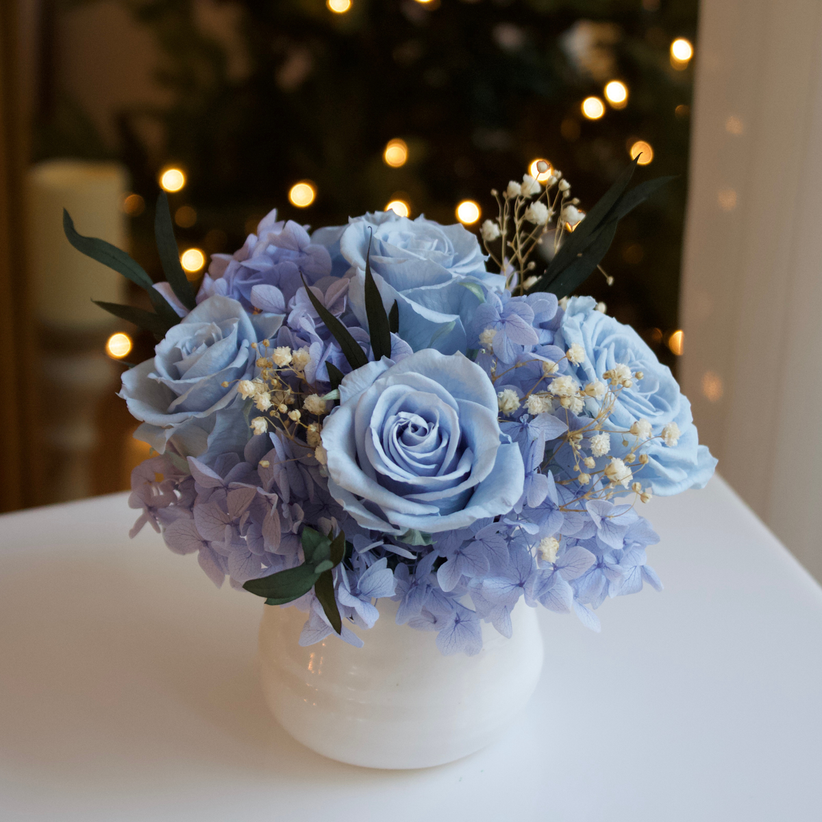 Mini by Eos Blooms: a white vase with blue hydrangeas and Forever Roses, softly lit behind.