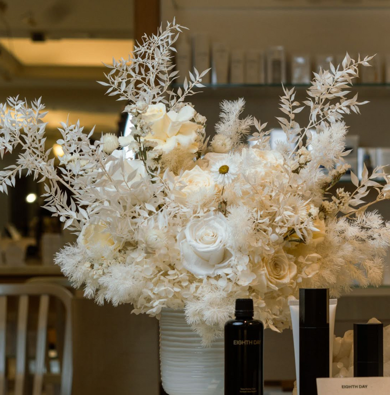 Elegant white floral arrangement in a vase, with black bottles and boxes in the foreground.