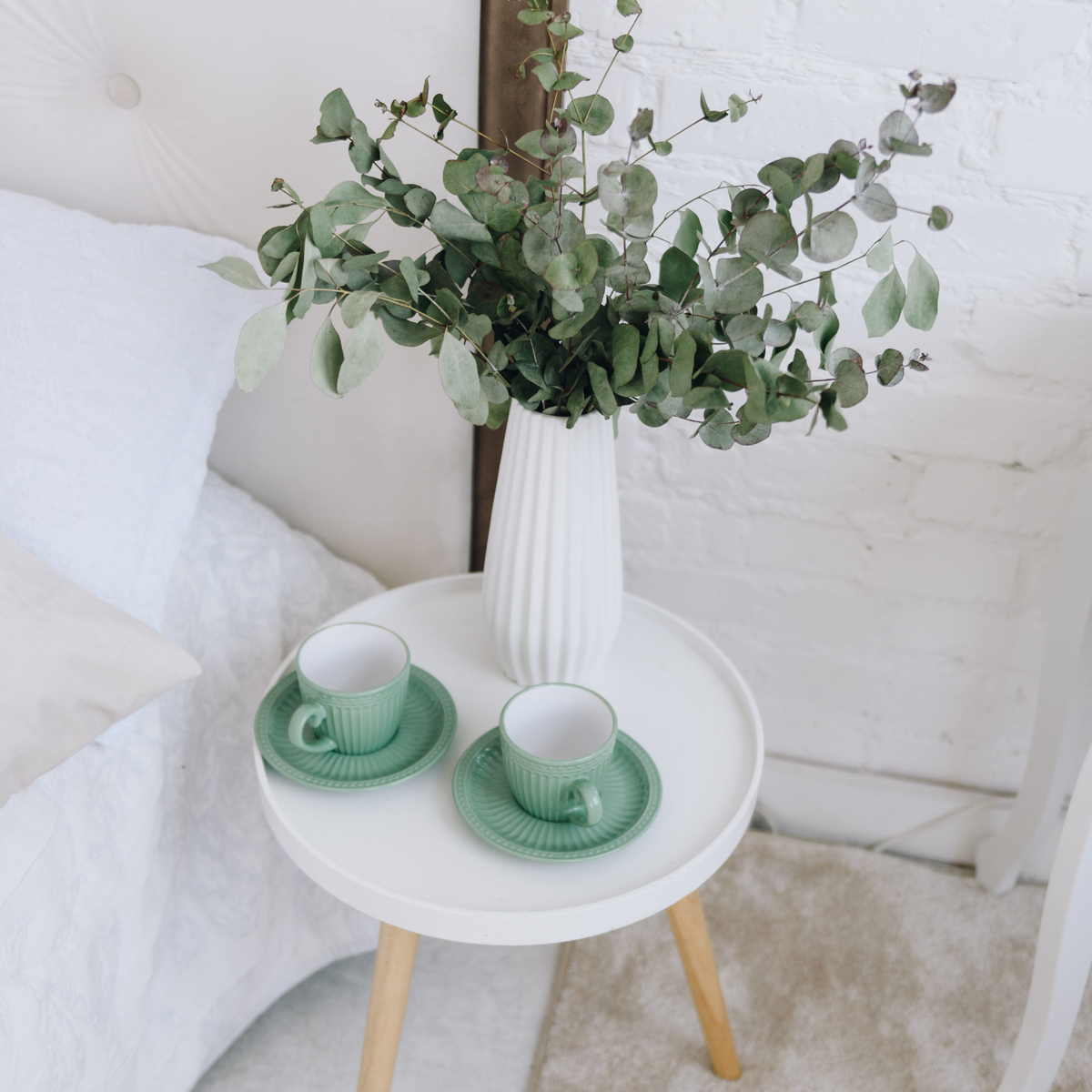 White bedside table with two green teacups and Eos Blooms Preserved Eucalyptus in a vase.
