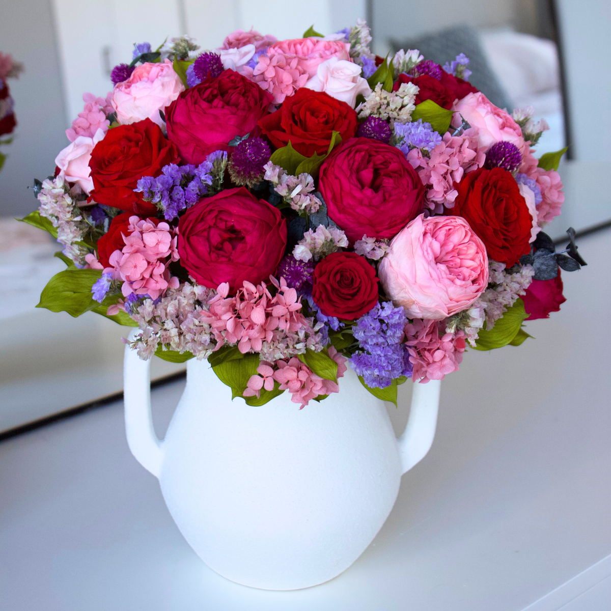 Bouquet of pink, red, and purple flowers in a white vase on a white surface.
