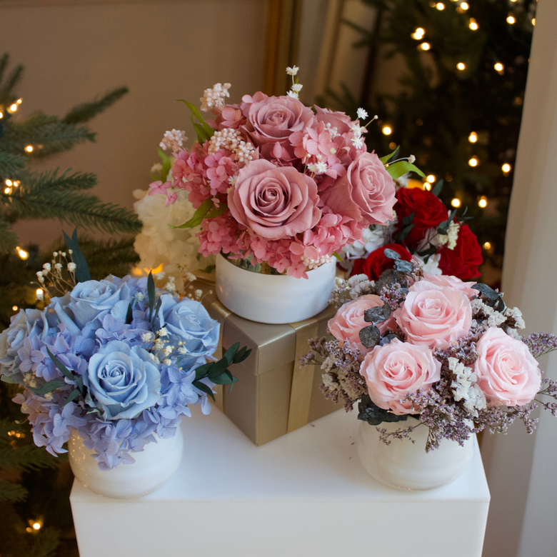 Three flower arrangements with pink, blue, and red roses on white stands, with festive lights.