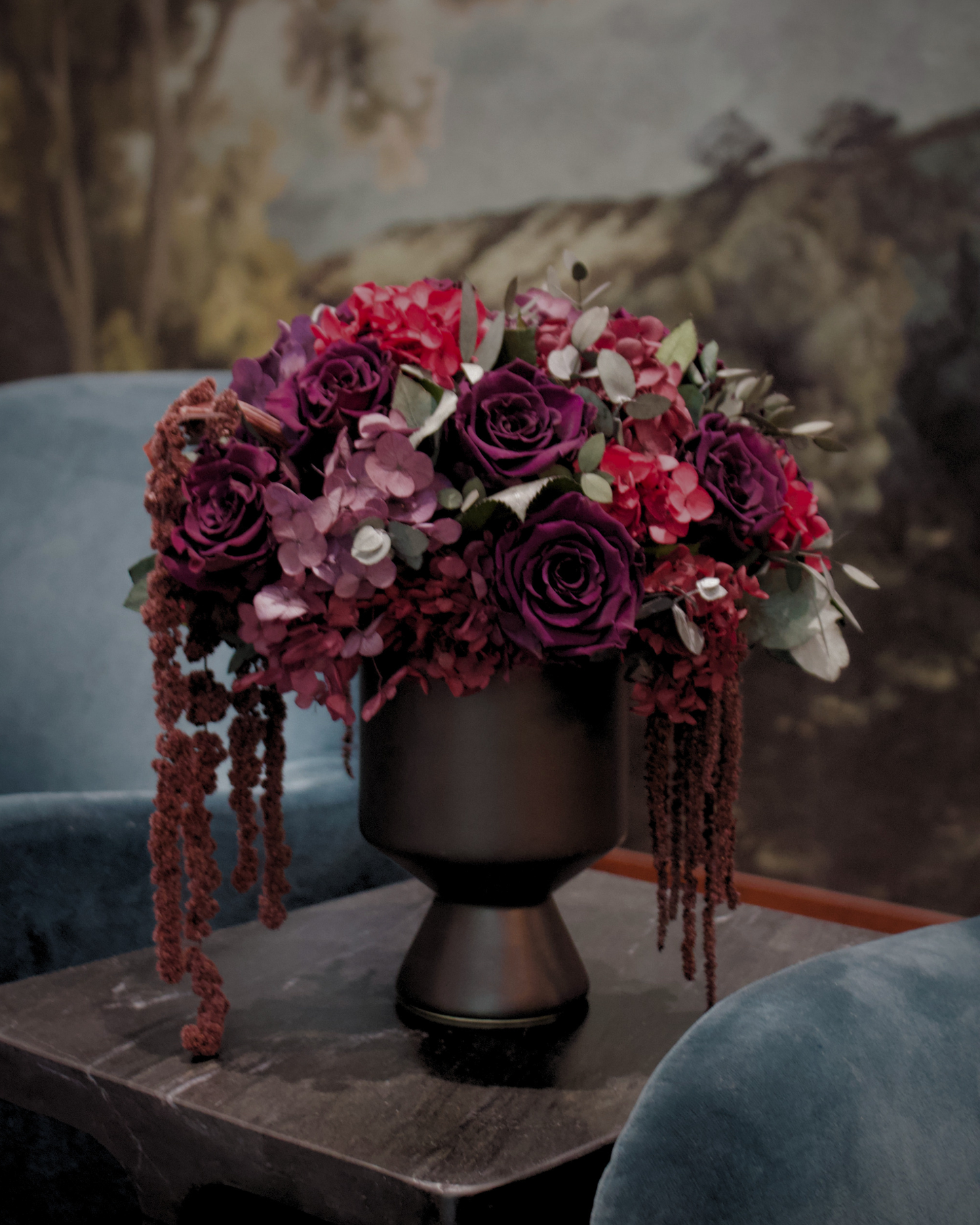 Eos Blooms’ After Hours vase with luxe florals rests on a dark table between blue chairs.