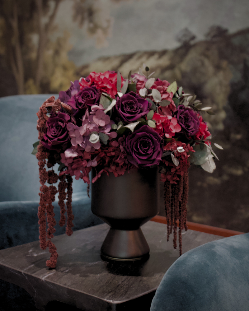 Eos Blooms’ After Hours vase with luxe florals rests on a dark table between blue chairs.