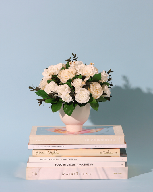 The Snowdrop (Petite) by Eos Blooms rests on books, set against a light blue background.