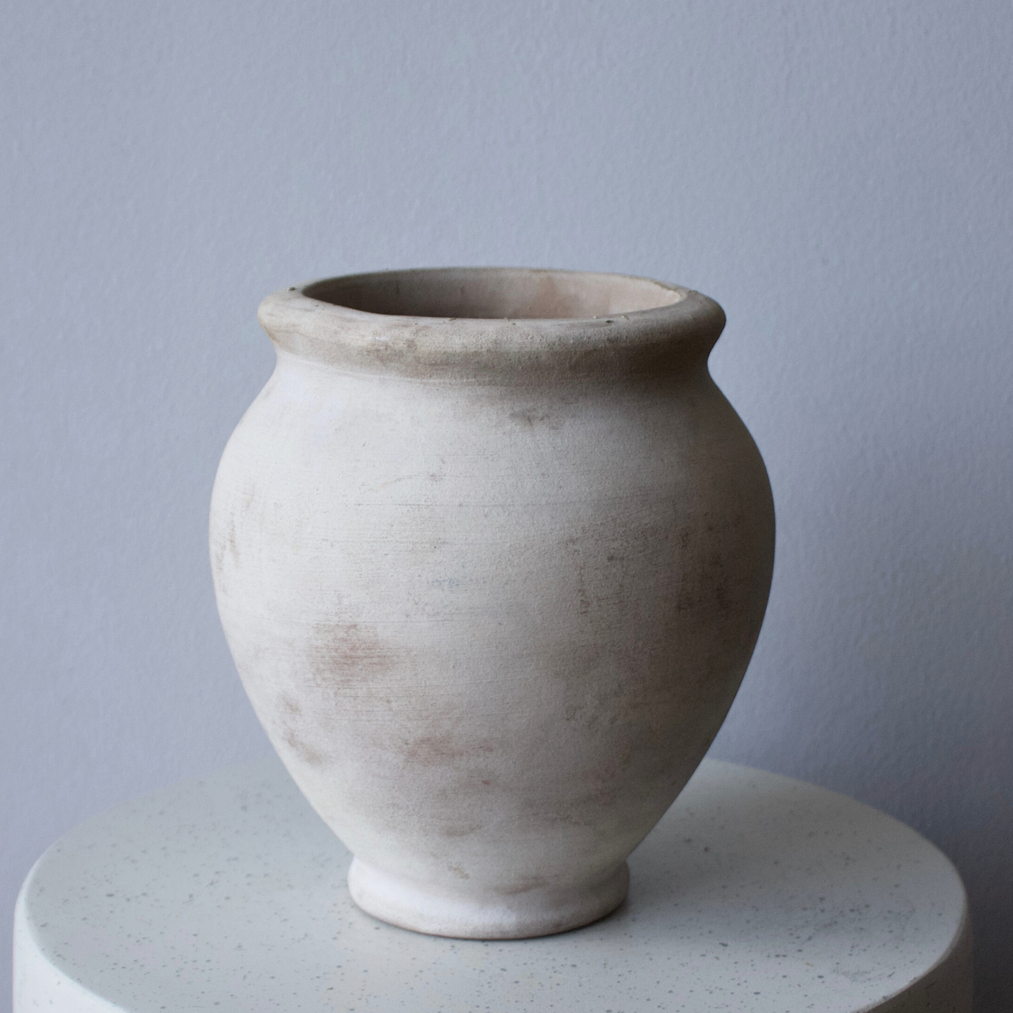 The Petra Vase by Eos Blooms, round and light clay, sits on white against a gray wall.