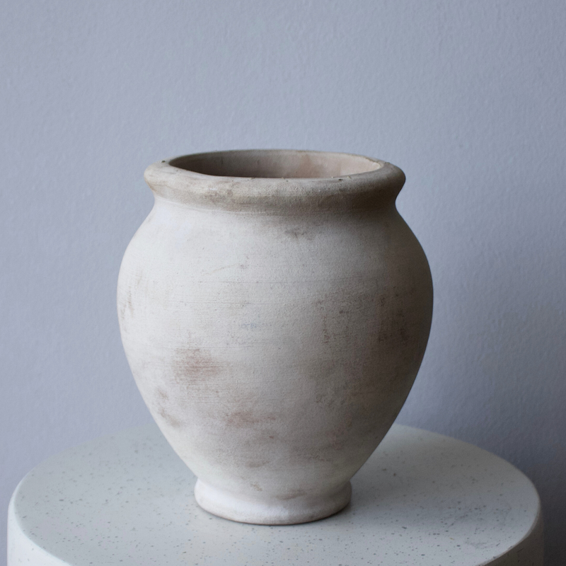 The Petra Vase by Eos Blooms, round and light clay, sits on white against a gray wall.
