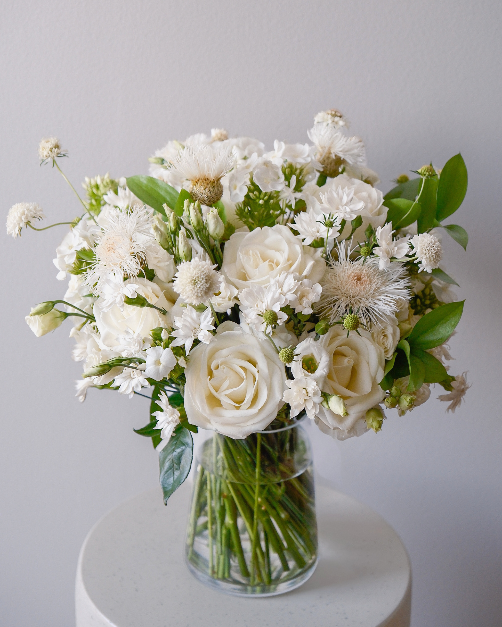 The Blanche by Eos Blooms: ivory garden roses with green leaves in a clear glass vase.