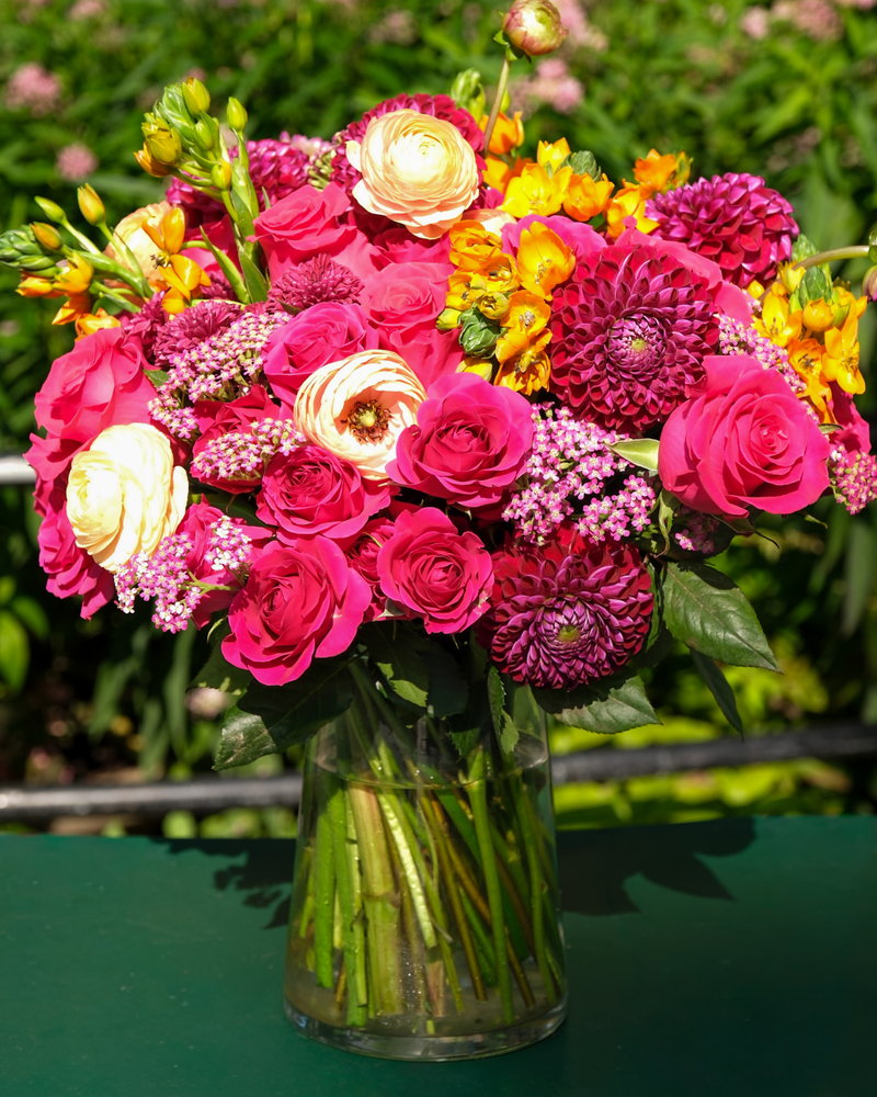 The Leo ♌ by Eos Blooms features pink, yellow, and purple blooms on a green outdoor table.