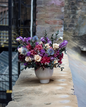 The Vinter (Petite) by Eos Blooms rests on stone before a rustic brick wall.