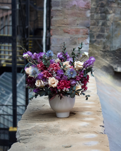 The Vinter (Petite) by Eos Blooms rests on stone before a rustic brick wall.
