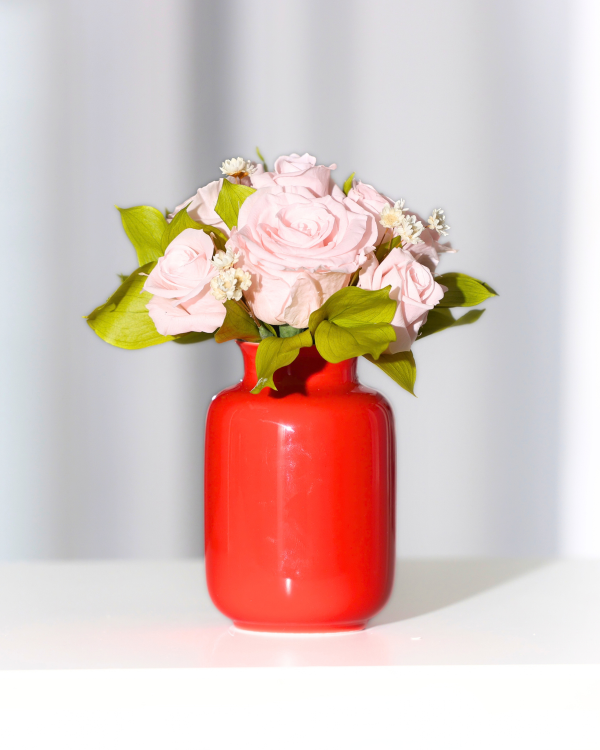 Eos Blooms Mini Milk Jars with forever roses and greenery in a red vase on white surface.