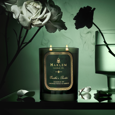 A lit Harlem candle beside white roses and a glass of water, against a dark green background.