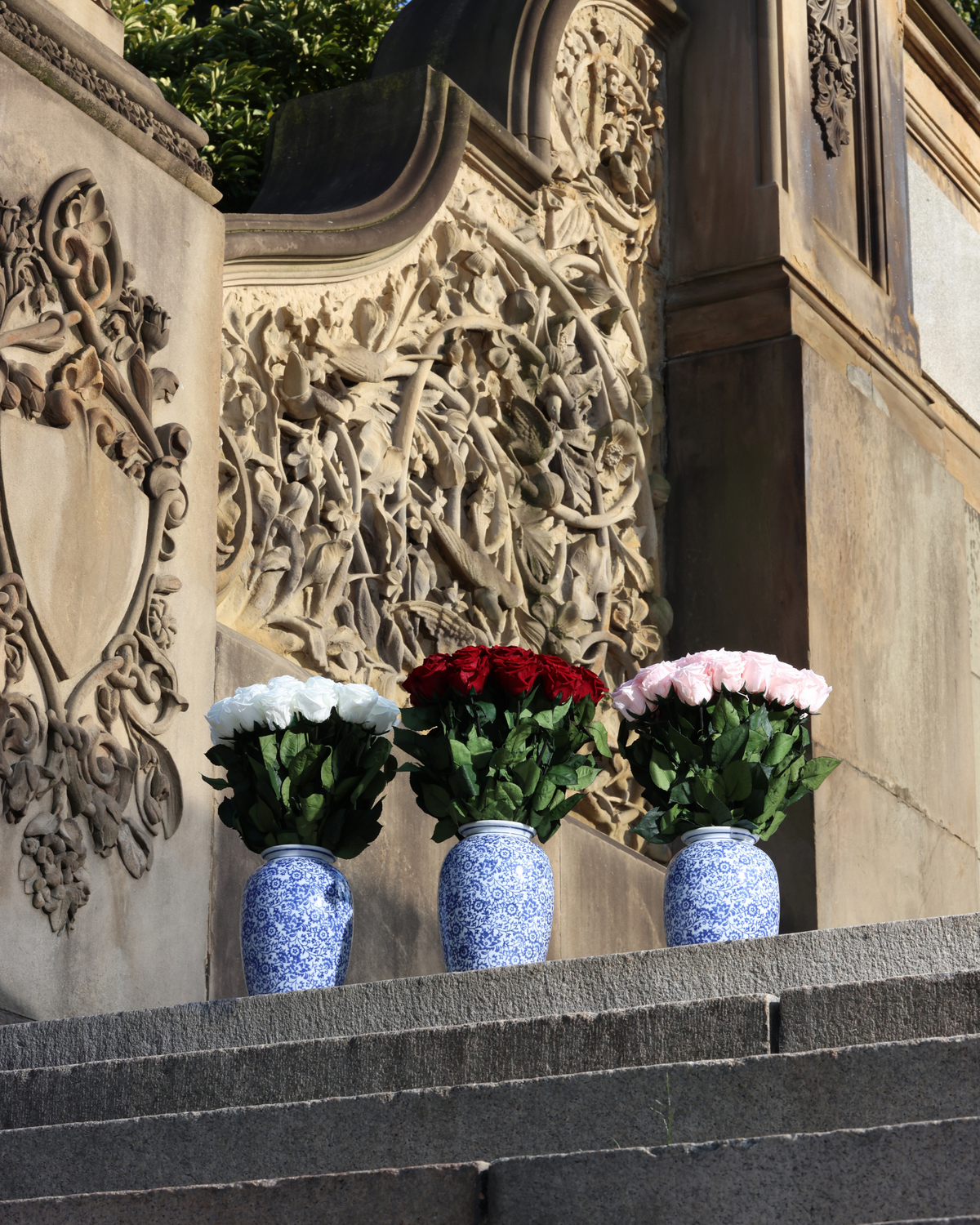 Three Eos Blooms Amalia vases with Forever Flowers sit on a stone ledge by ornate carvings.