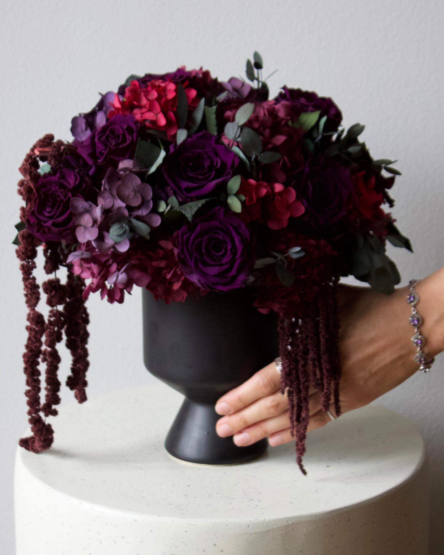 A hand arranges Eos Blooms’ After Hours, a sculptural dark floral bouquet, on a white table.