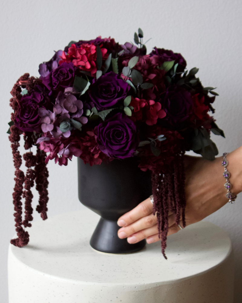 A hand arranges Eos Blooms’ After Hours, a sculptural dark floral bouquet, on a white table.