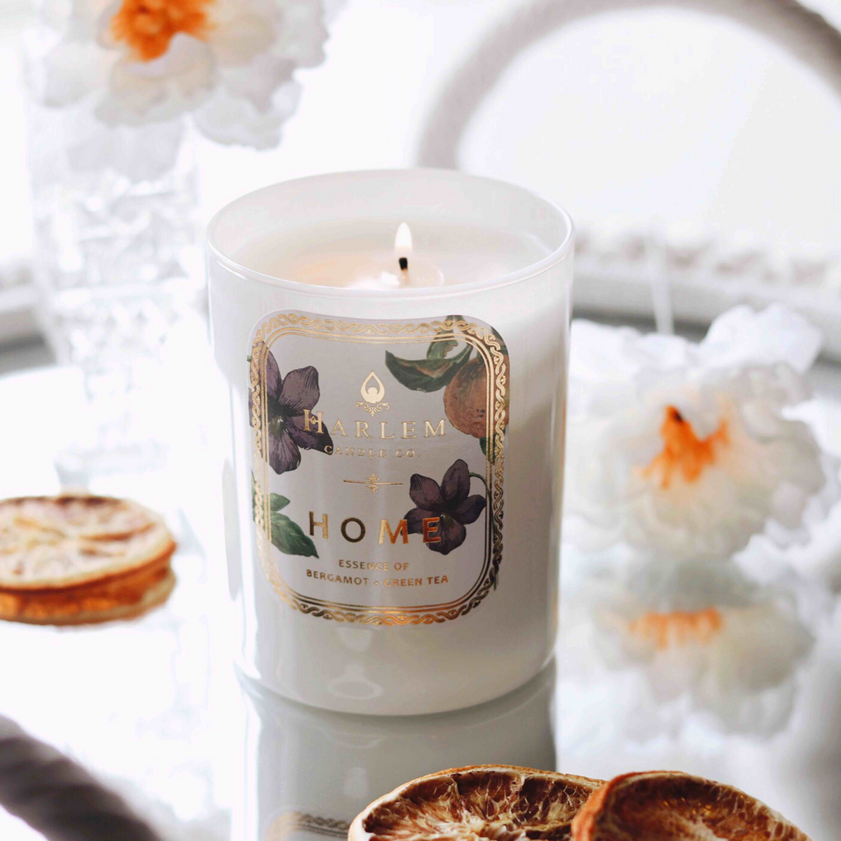 White candle labeled Home burning on a reflective surface with flowers and dried citrus.