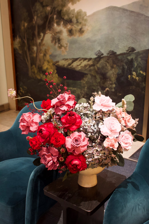 A vase of pink and red flowers sits on a table between two blue chairs.