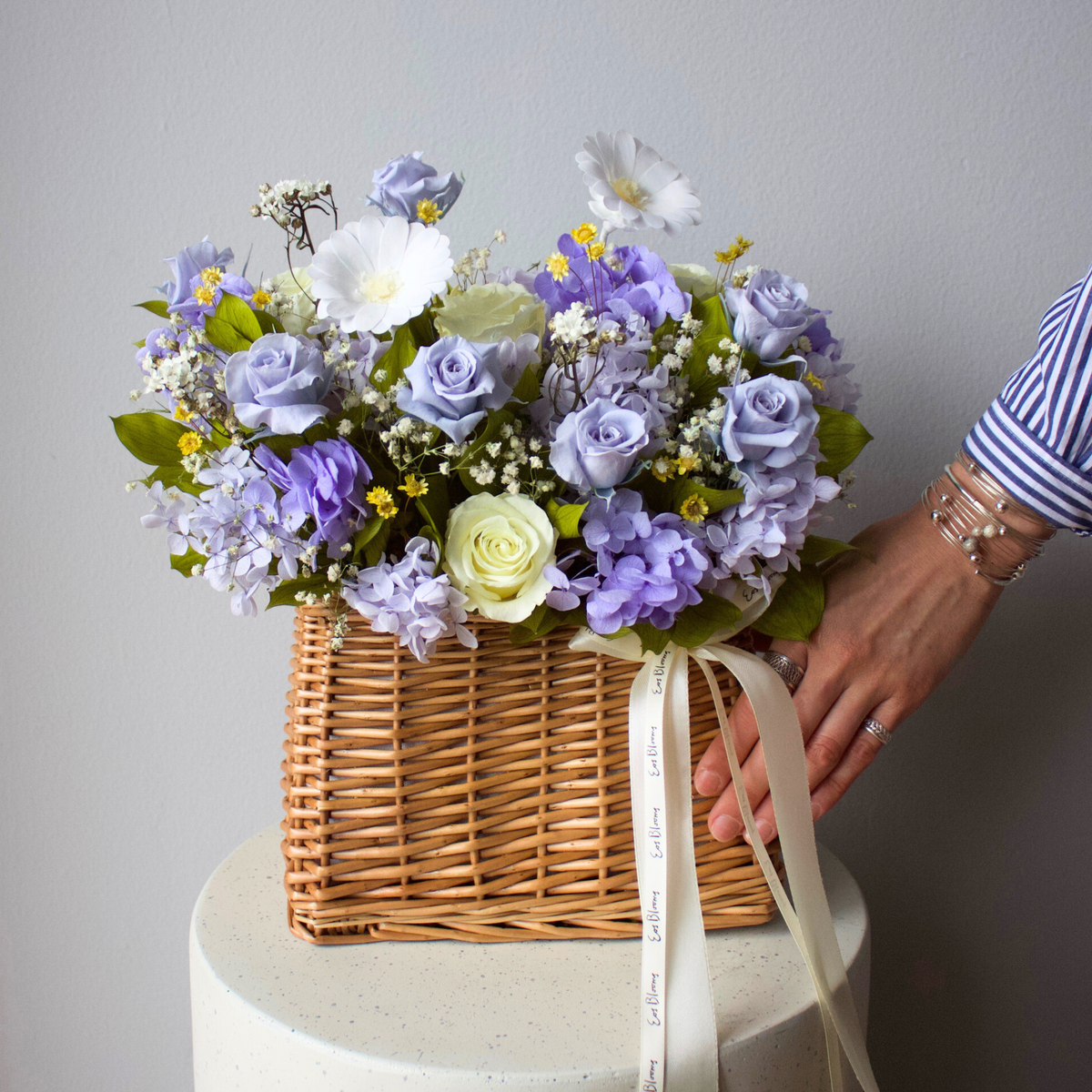 A wicker basket with pastel flowers, mostly purple and white, with a hand adjusting a ribbon.