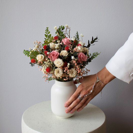 A hand touches a vase of mixed pink and white flowers on a round white table.