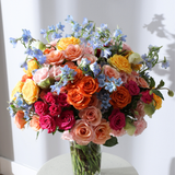 A vibrant bouquet of mixed roses and blue flowers in a glass vase on a white table.