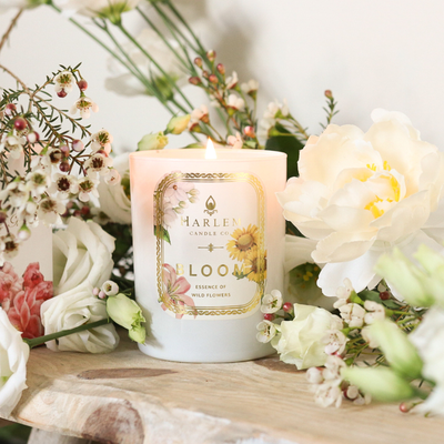 White candle labeled BLOOM surrounded by white and pink flowers on a wooden surface.