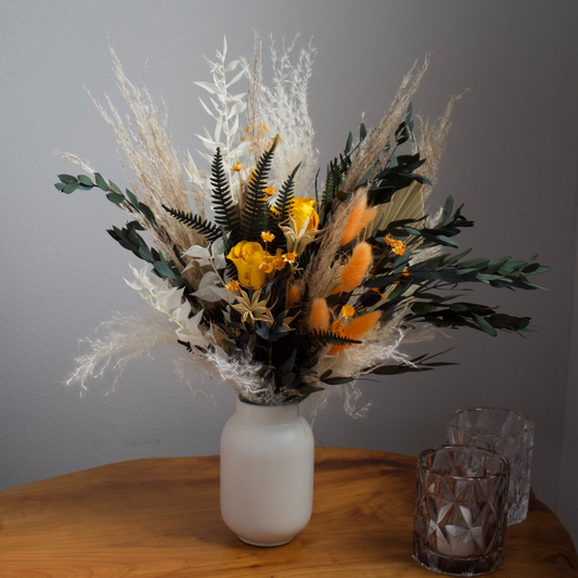 Harvest bouquet by Eos Blooms with yellow preserved flowers in a white vase on wood table.