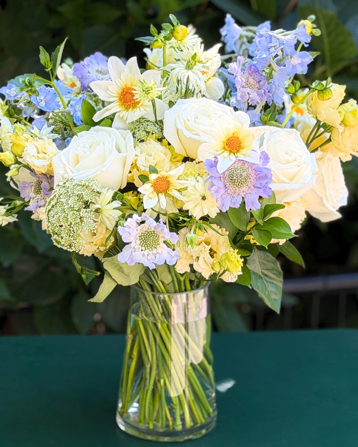 The Libra ♎ by Eos Blooms: white, yellow & light purple flowers on a green table.