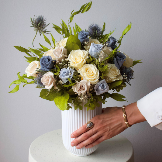 A hand arranges Eos Blooms’ Preserved Flower Subscription of white and blue roses on a table.