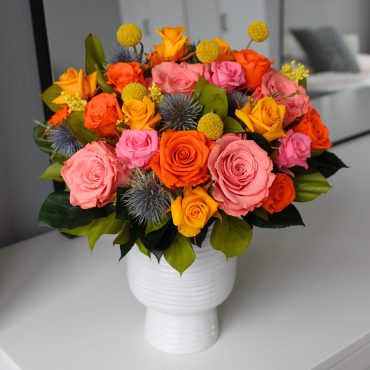 A vibrant bouquet of pink, orange, and yellow roses in a white vase on a table.